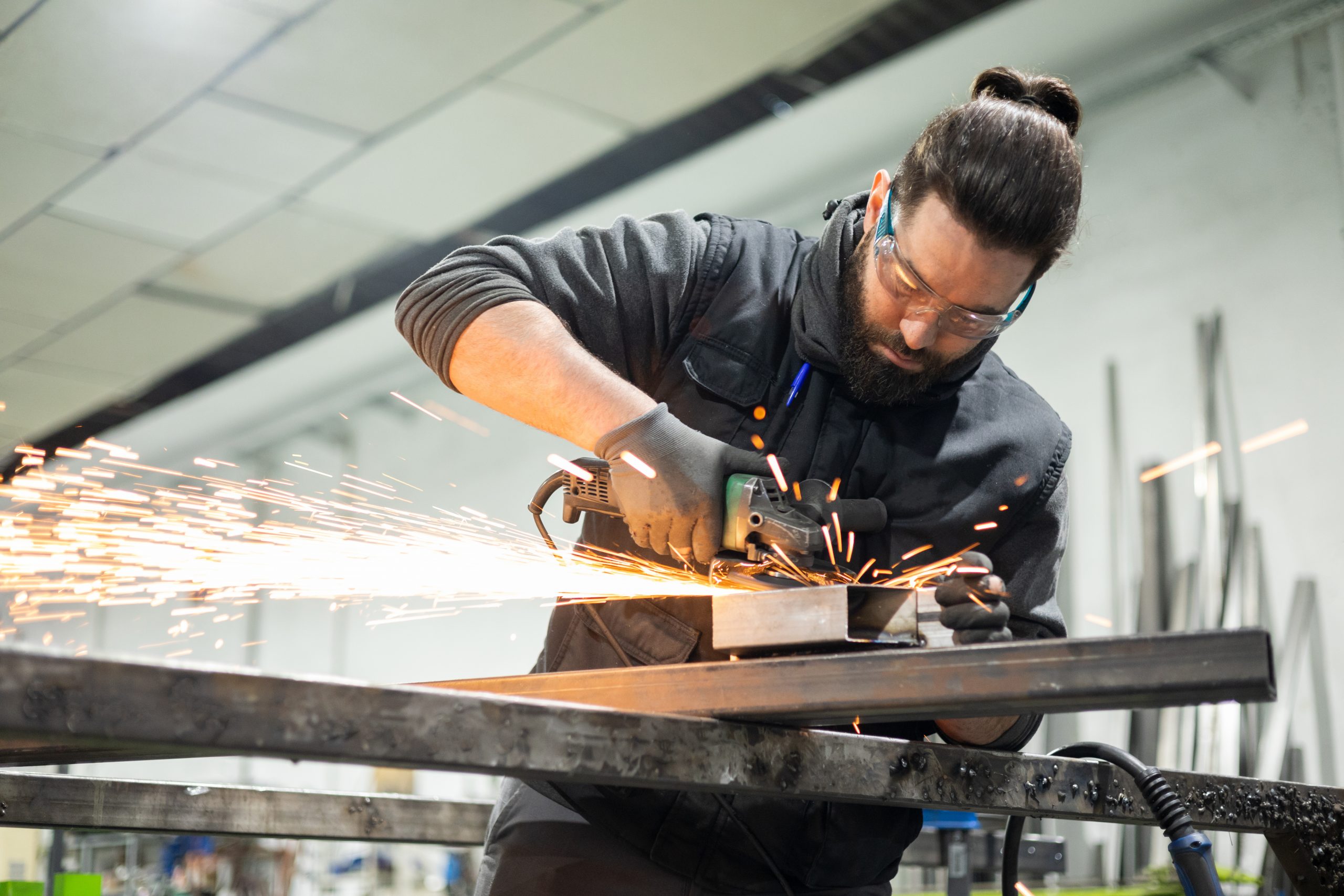Ein Mann in Schutzkleidung und mit Brille benutzt eine Radialschleifmaschine, um in der Werkstatt ein Metallprofil zu schneiden.